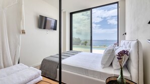 A modern bedroom with a large window, a bed, a TV, and a view of the sea. - Alpha VIlla | S Cave Villas Seaside Karystos (Karystos)