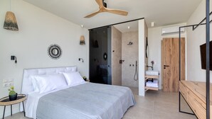 A modern bedroom with a large bed, a ceiling fan, a shower area, and a wooden vanity.