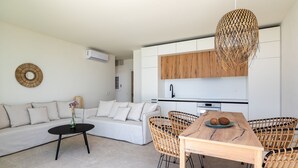 A modern living room with a sofa, coffee table, and wicker chairs. A wooden dining table with a plate of nuts is in the center.