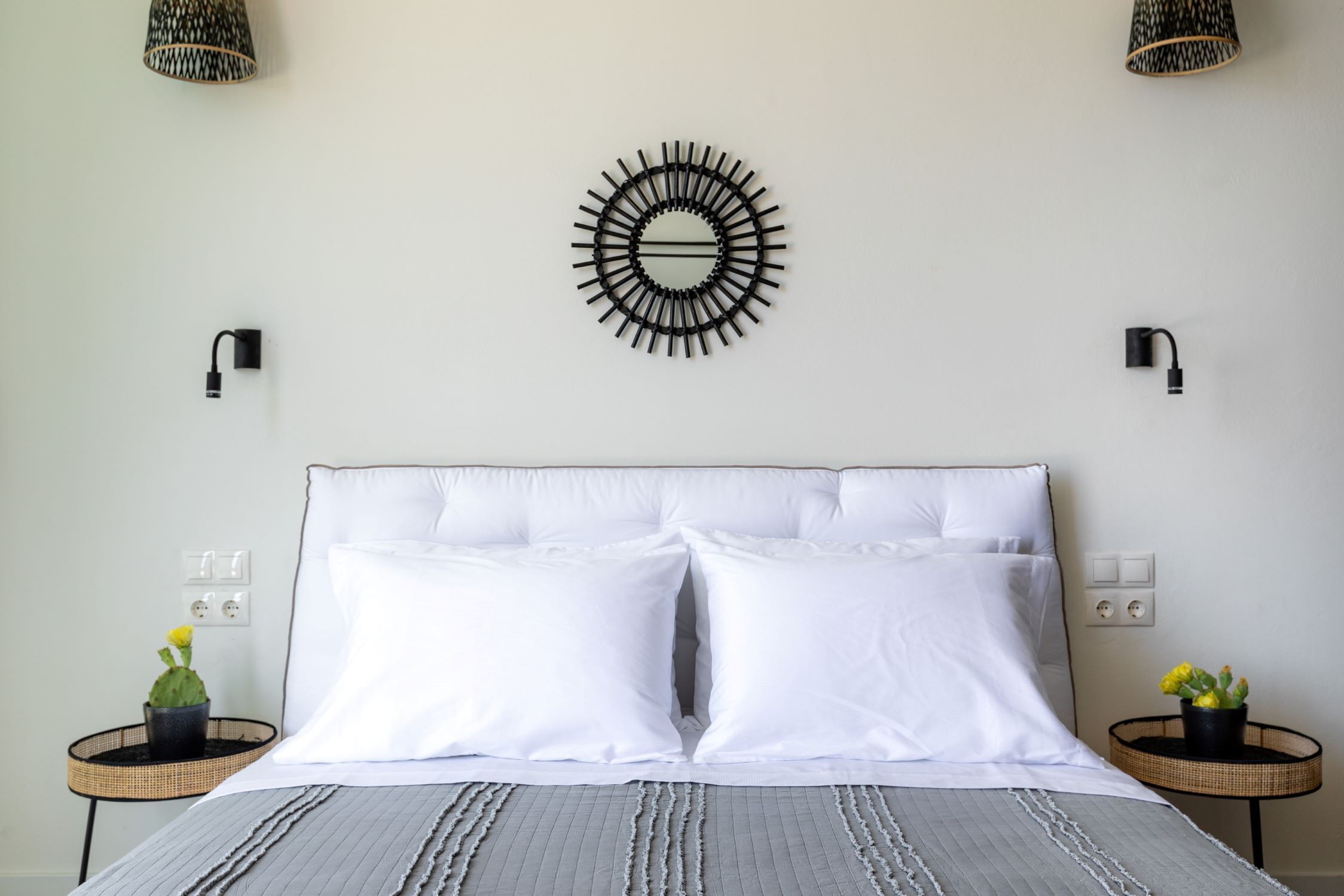 A neatly made bed with white bedding, two bedside tables with potted plants, a wall-mounted circular light fixture, and two wall-mounted light switches.