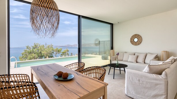 A modern living room with a wooden table, wicker chairs, a sofa, and a view of the sea.
