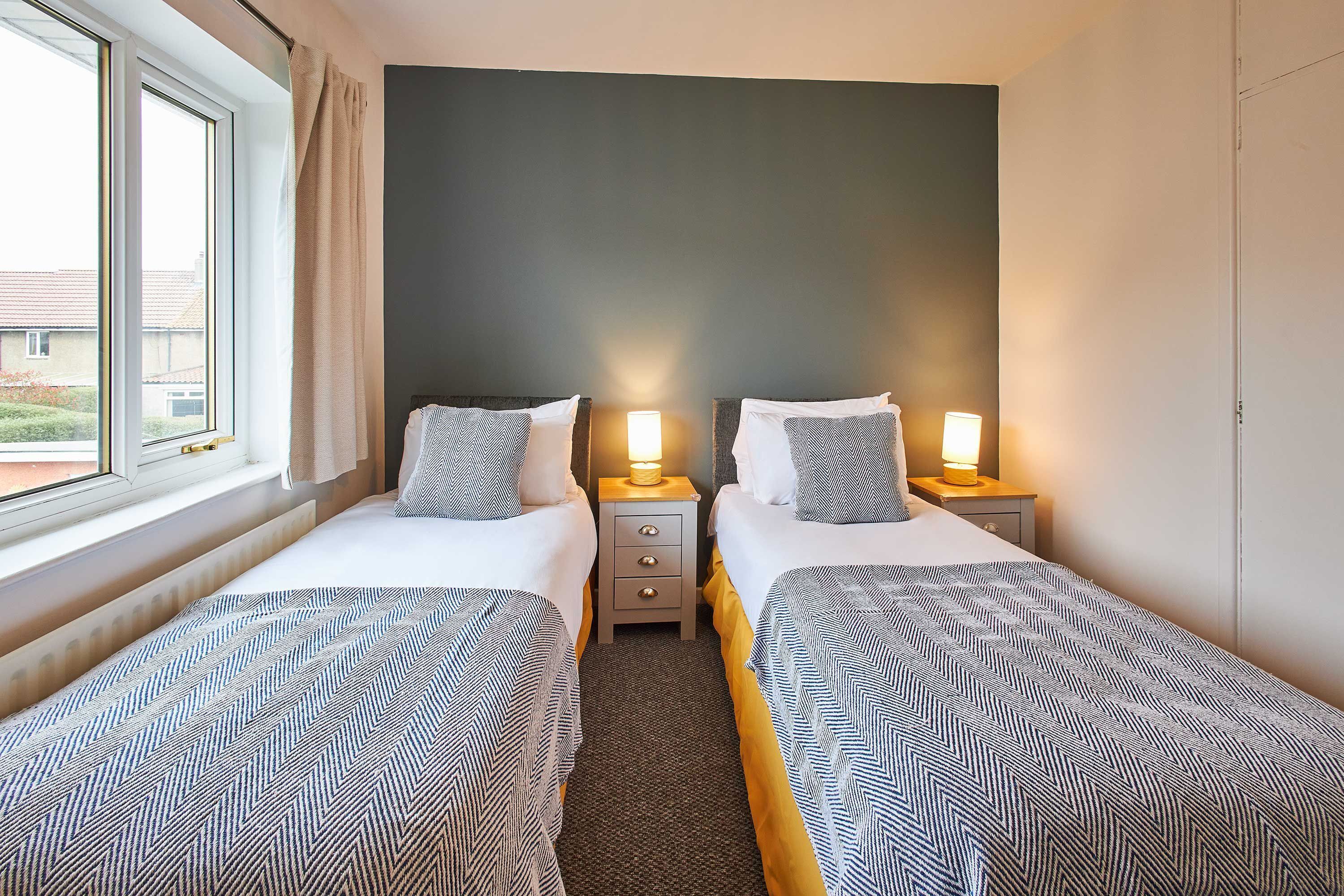A hotel room with two single beds, a window, a nightstand with a lamp, and a dark wall.