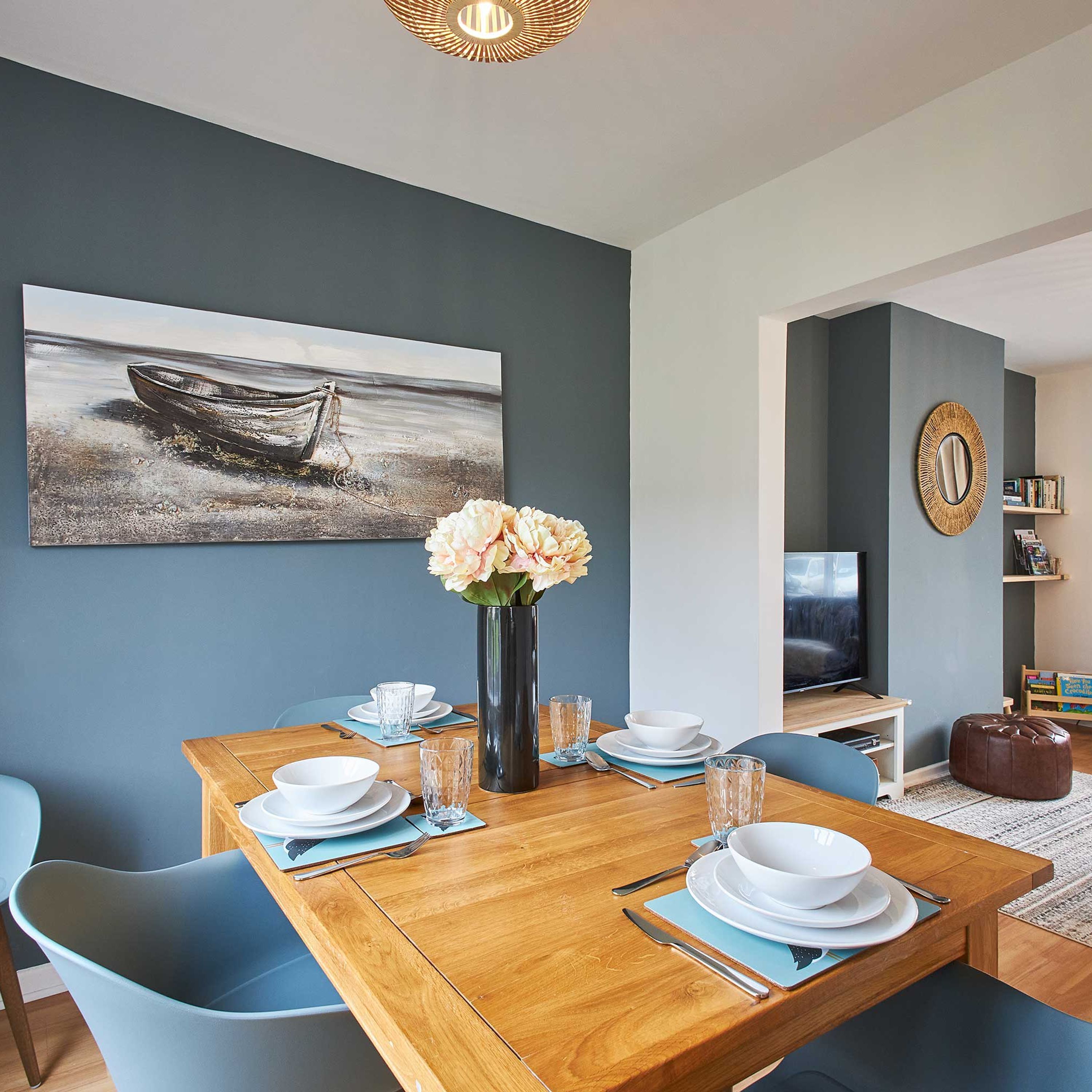 A dining area with a wooden table set for four, blue chairs, a vase with flowers, and a painting of a boat on the wall.