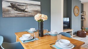 Un comedor con una mesa de madera preparada para cuatro, sillas azules, un florero con flores y un cuadro de un barco en la pared.