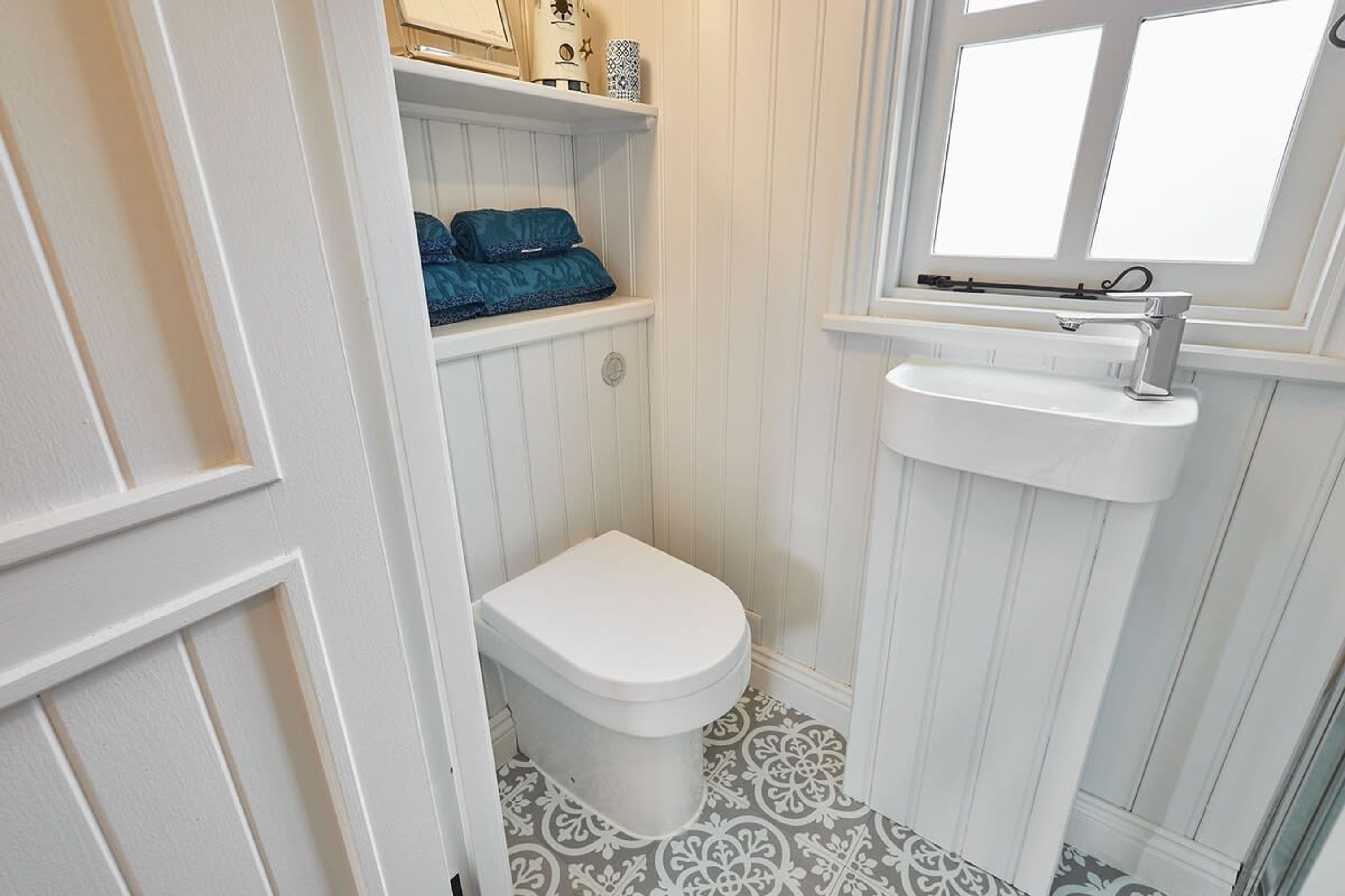 Ein kleines, weißes Badezimmer mit Toilette, einem Regal mit einem blauen Gegenstand und einem Fenster über einem Waschbecken.