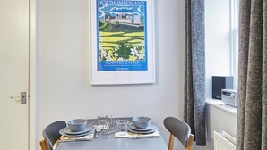 Un coin repas avec une table dressée pour quatre personnes, des chaises, un tableau encadré représentant le Northumberland et une fenêtre avec des rideaux.