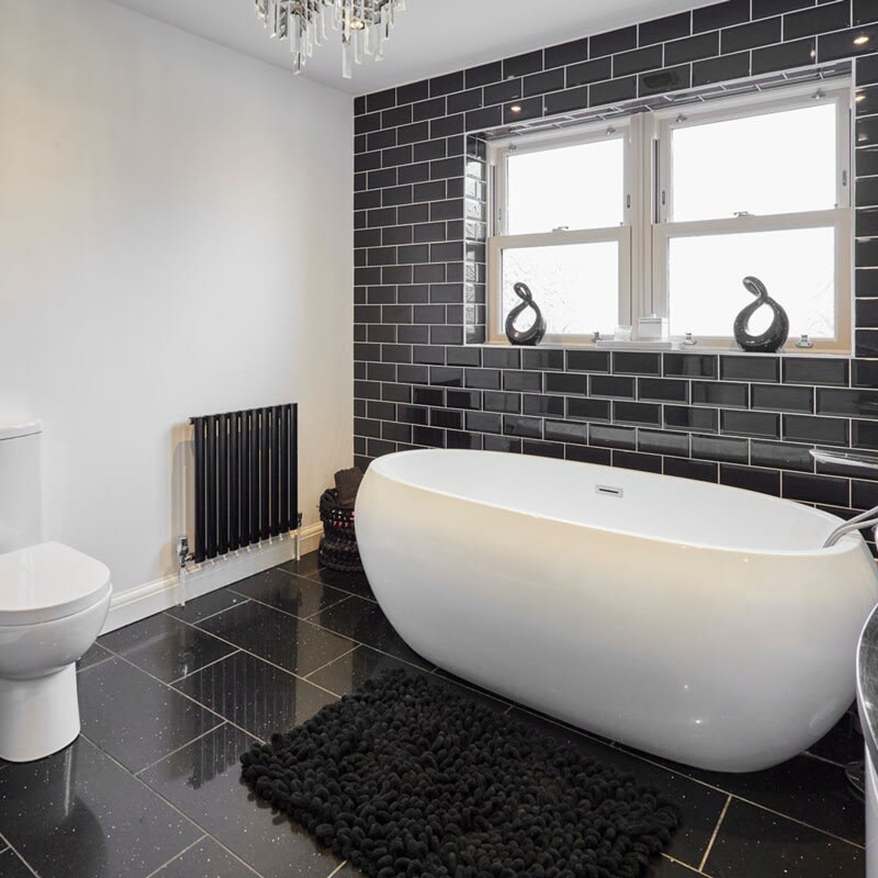 A modern bathroom with a freestanding bathtub, black tiled walls, and a window with decorative items.