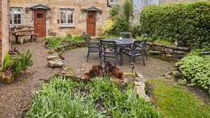 A stone house with a garden patio, a wooden bench, and a table with chairs.