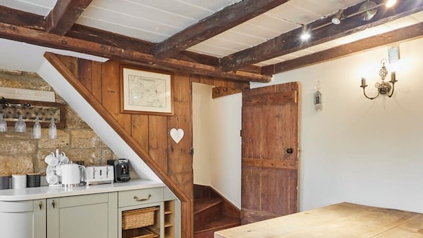 A rustic kitchen with a wooden table, a staircase, and a stone wall with jars.