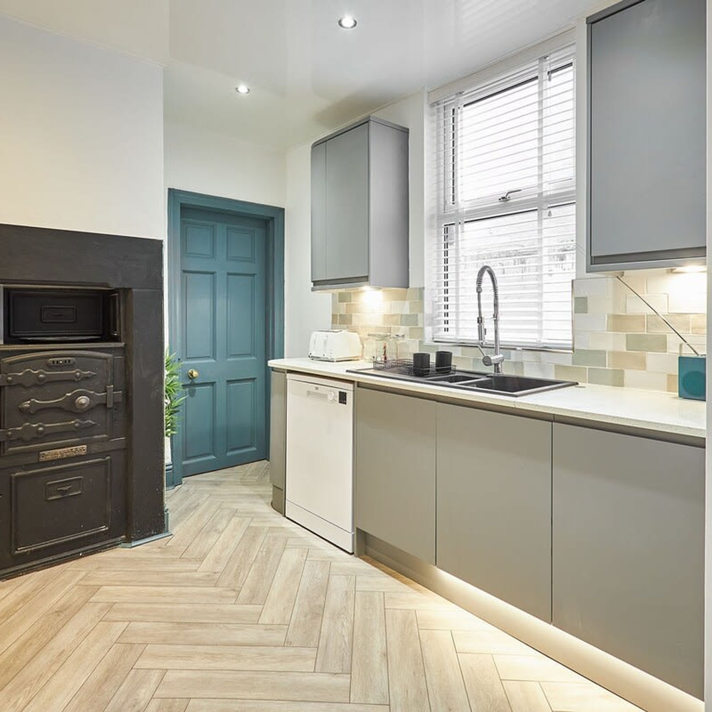 A compact kitchen with a herringbone floor pattern, a built-in dishwasher, and a window with blinds.