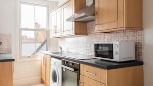 A compact kitchen with wooden cabinets, a washing machine, a microwave, and a window with a view of buildings. - Host Stay Abbey View (Whitby)