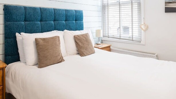 A bedroom with a blue upholstered headboard, white bedding, and a window with blinds. - Host Stay Abbey View (Whitby)