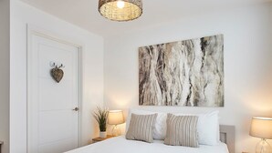 A neatly made bed with white linens and striped pillows, a white door with a heart-shaped decoration, a wall-mounted abstract painting, and two bedside tables with lamps. - Host Stay Emilyn House (Sunderland)