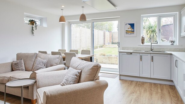 A modern living room with a sofa, a small table, and a kitchen area with cabinets and a window. - Host Stay Emilyn House (Sunderland)