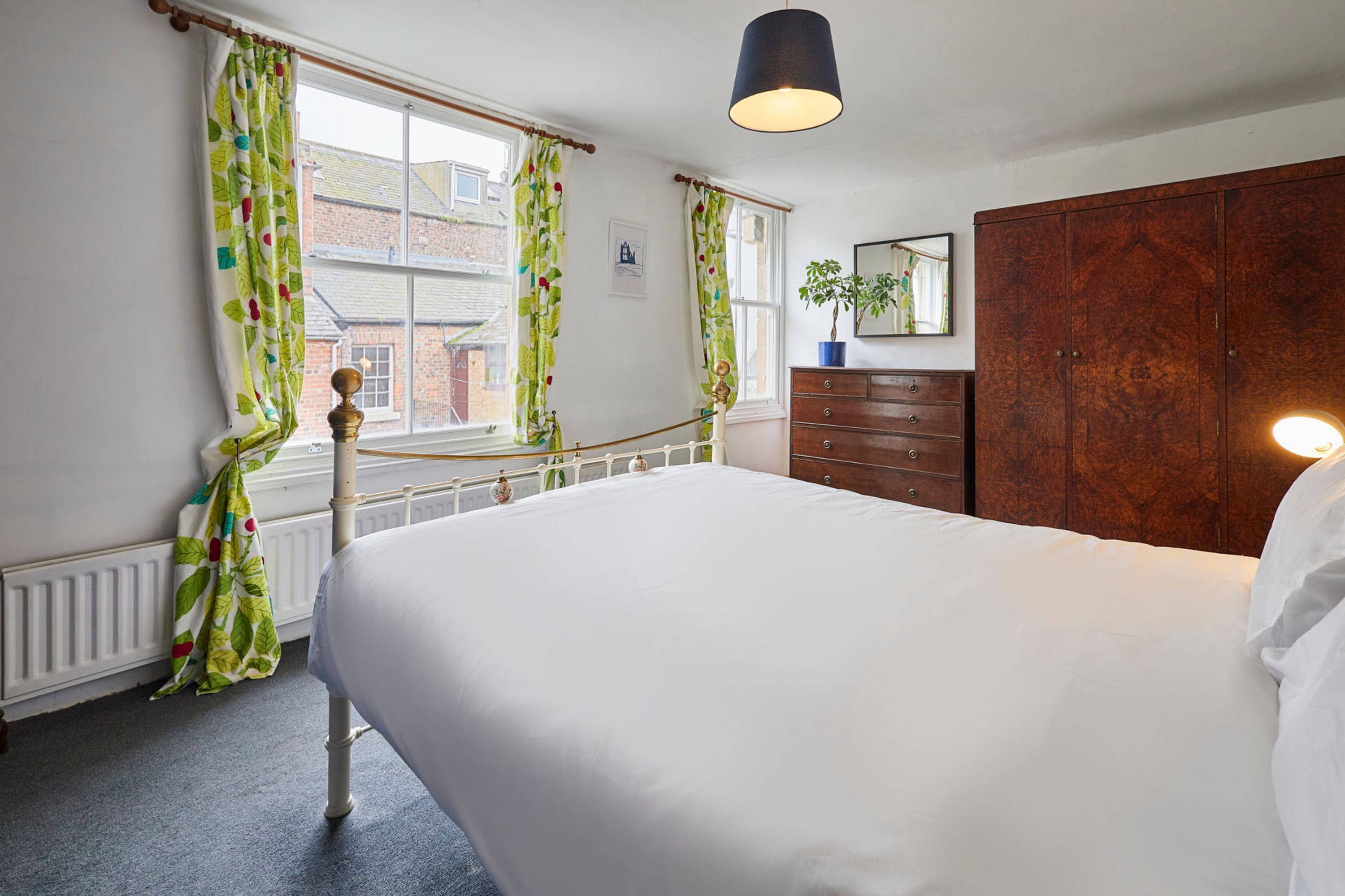 A bedroom with a large bed, a wooden wardrobe, a dresser, a mirror, and a window with curtains.