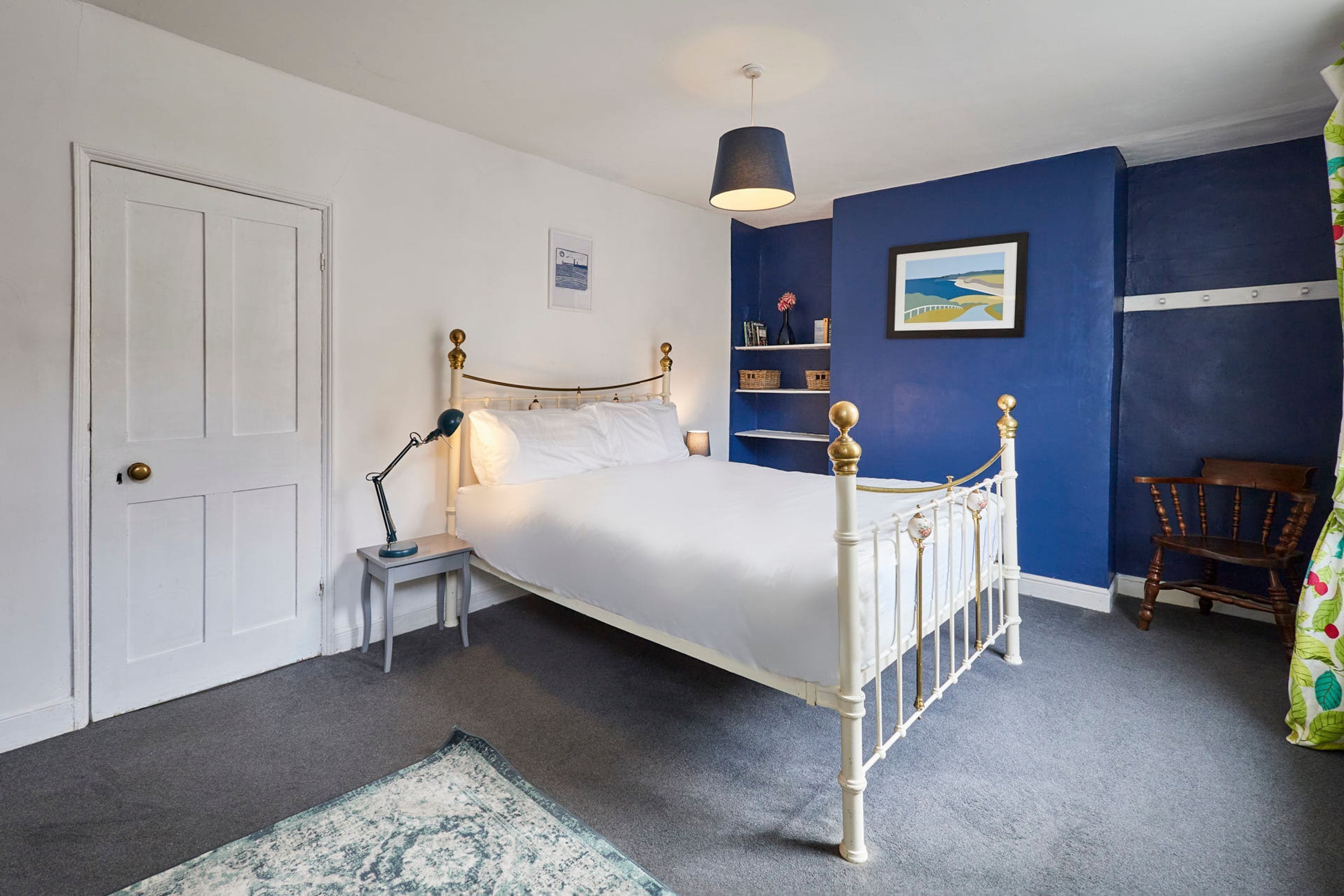 Een slaapkamer met een wit bed, een bedlampje, een nachtkastje, een blauwe wand, een houten stoel en een deur.