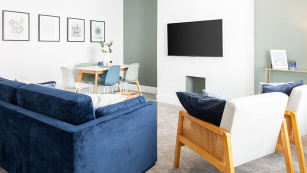 Un salon moderne avec un canapé bleu, une chaise en bois, une table à manger avec des chaises, un téléviseur à écran plat fixé au mur et des œuvres d’art encadrées accrochées au mur.