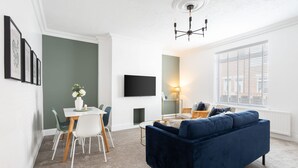 Un salon moderne avec un canapé bleu foncé, une table à manger avec des chaises, un téléviseur à écran plat fixé au mur et une fenêtre avec des stores.