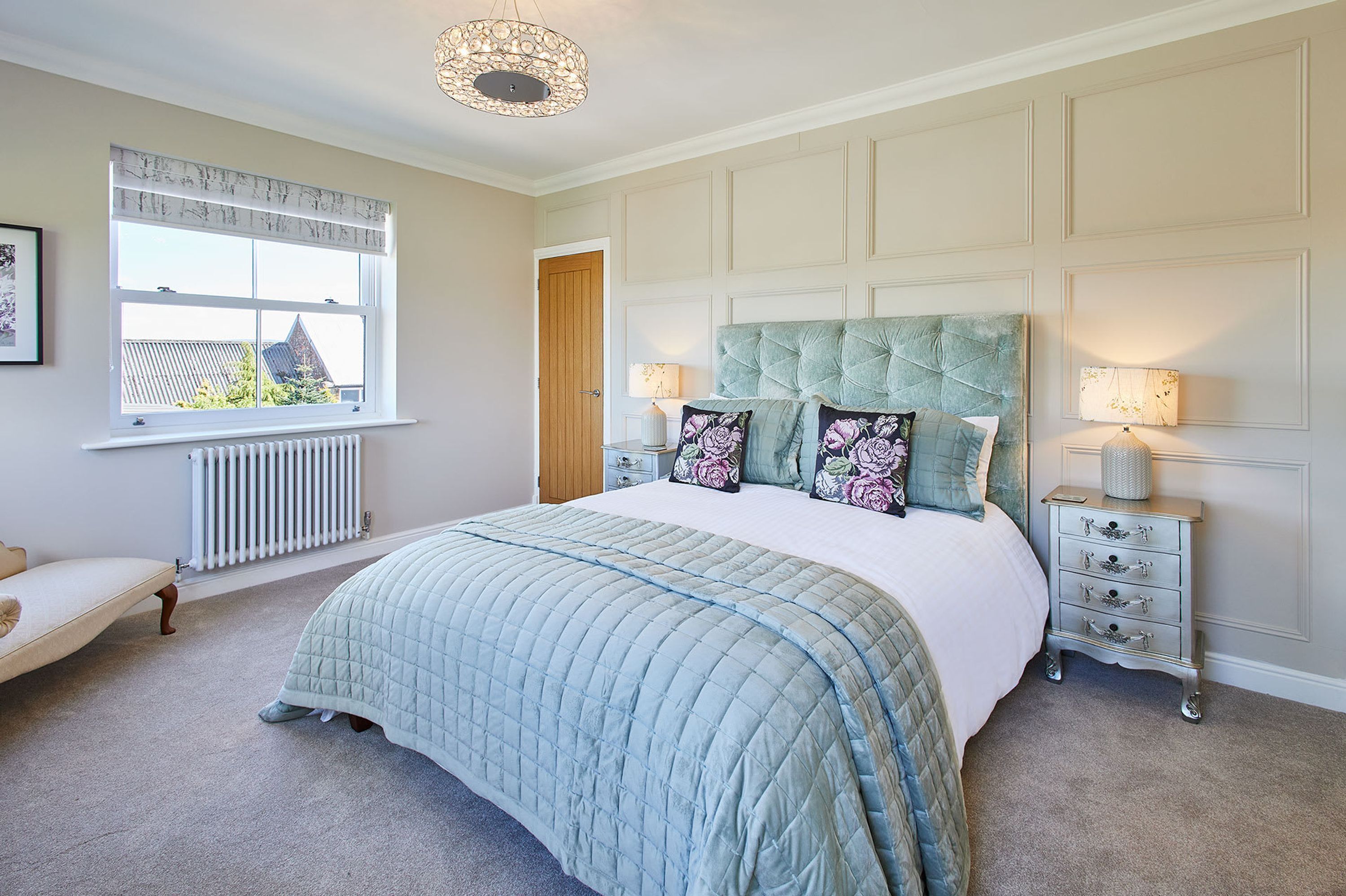Een slaapkamer met een groot bed, een nachtkastje, een lamp, een radiator, een raam met gordijnen en een deur.