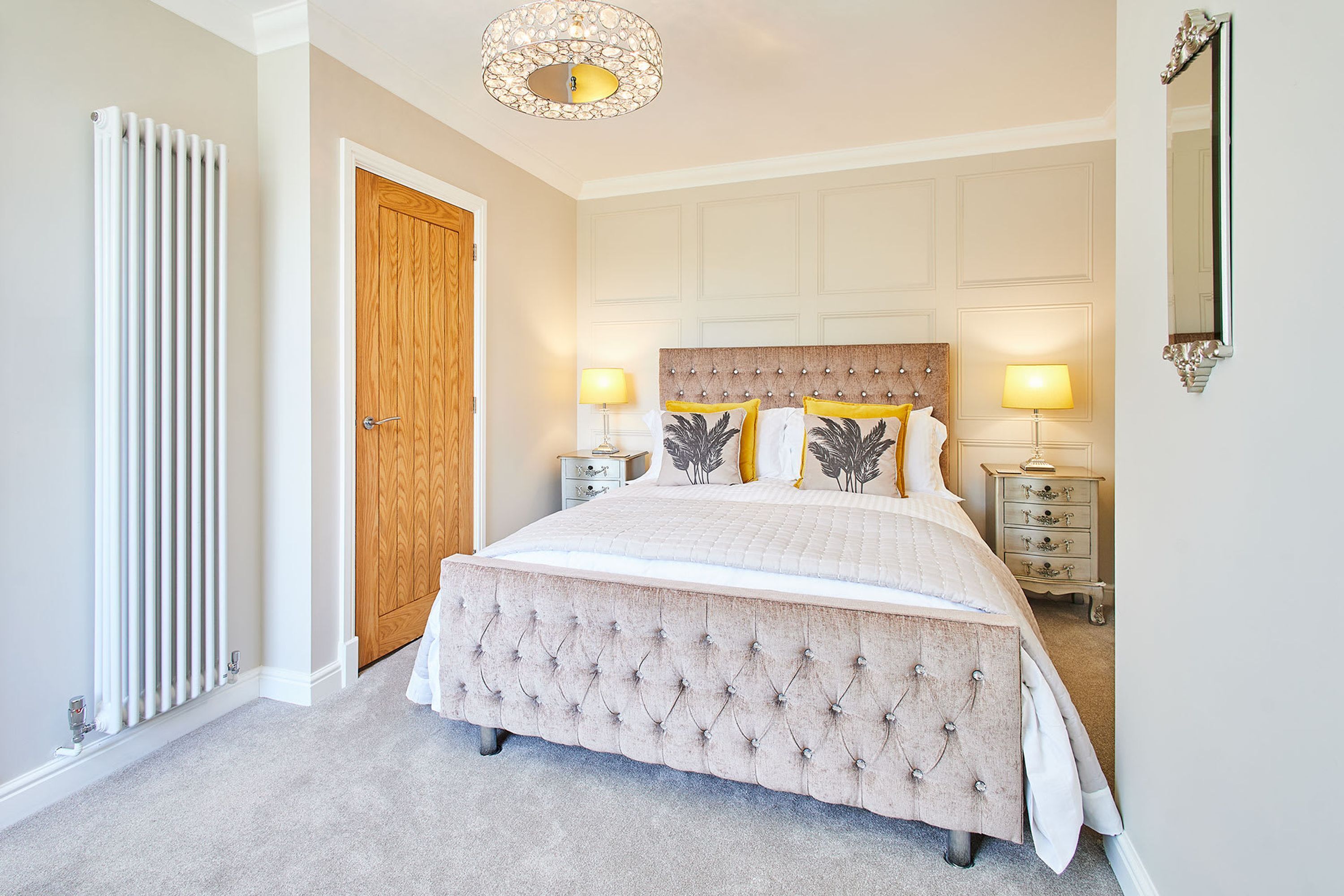 A bedroom with a large bed, two bedside tables, a wooden door, a radiator, and a chandelier.