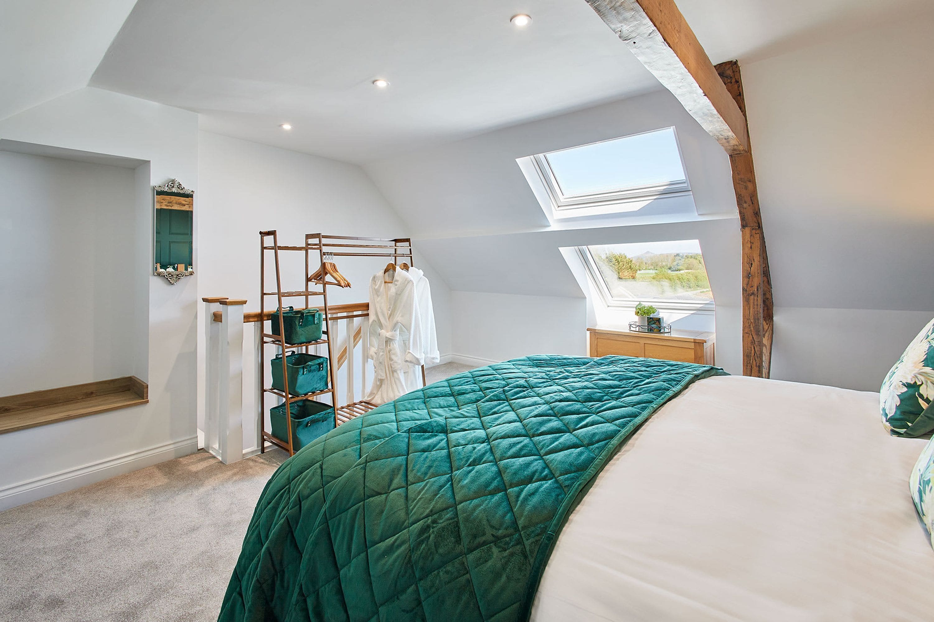 A bedroom with a skylight, a bed with a green quilt, a wooden wardrobe, and a hanging rack with clothes.