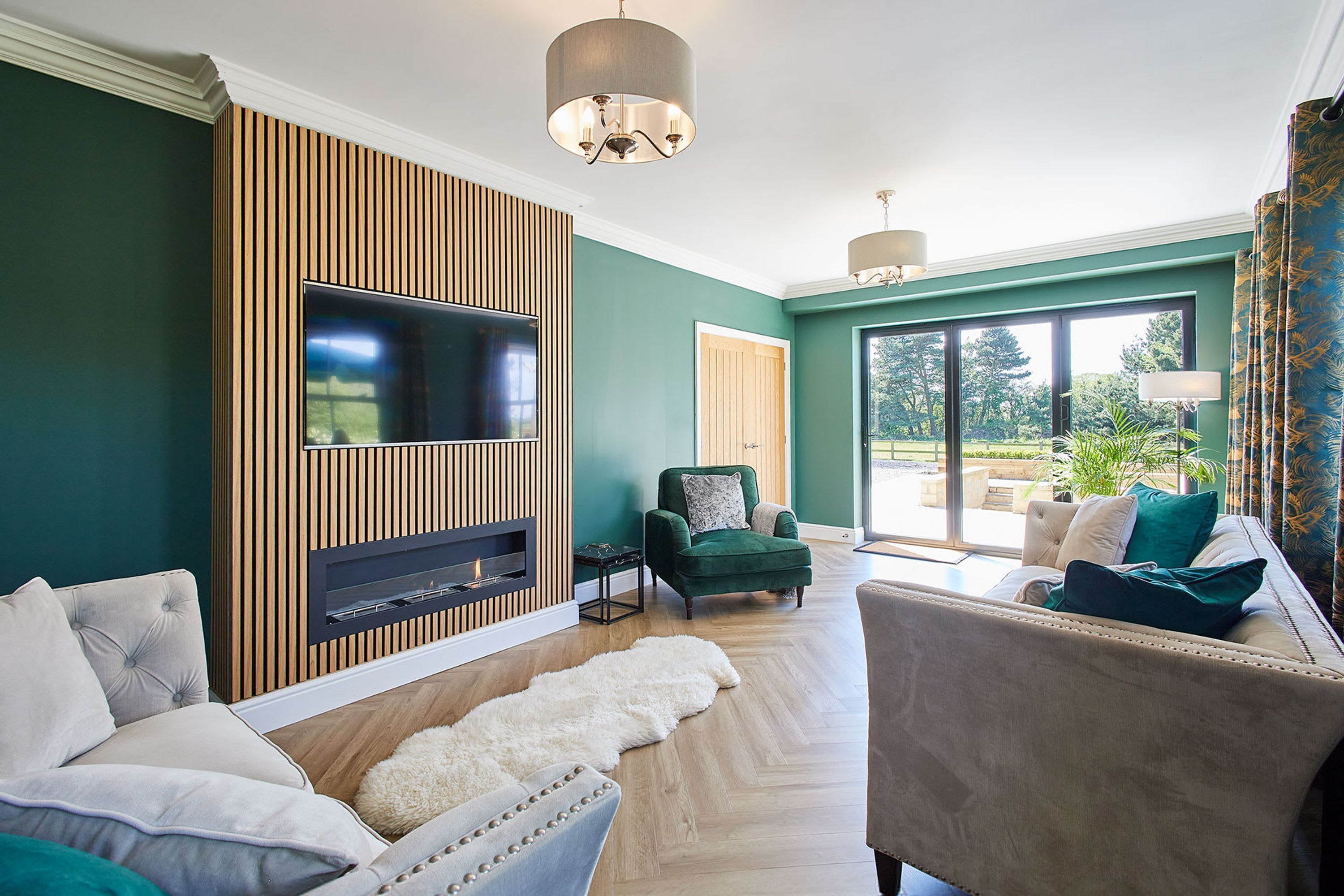 A living room with a TV, fireplace, green armchair, sofa, and a view of the outdoors.