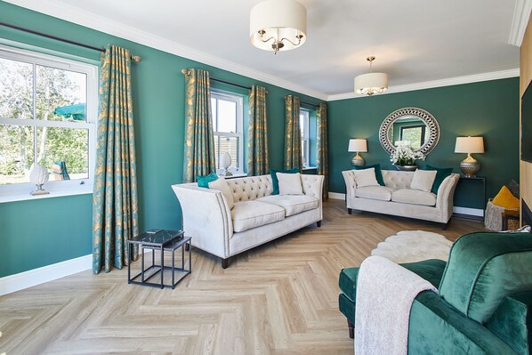 Una sala de estar con dos sofás blancos, un sillón verde, piso de madera con diseño espiga y un espejo grande en la pared.
