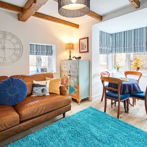 A cozy living room with a brown leather sofa, a teal rug, and a wooden dining table with blue chairs.