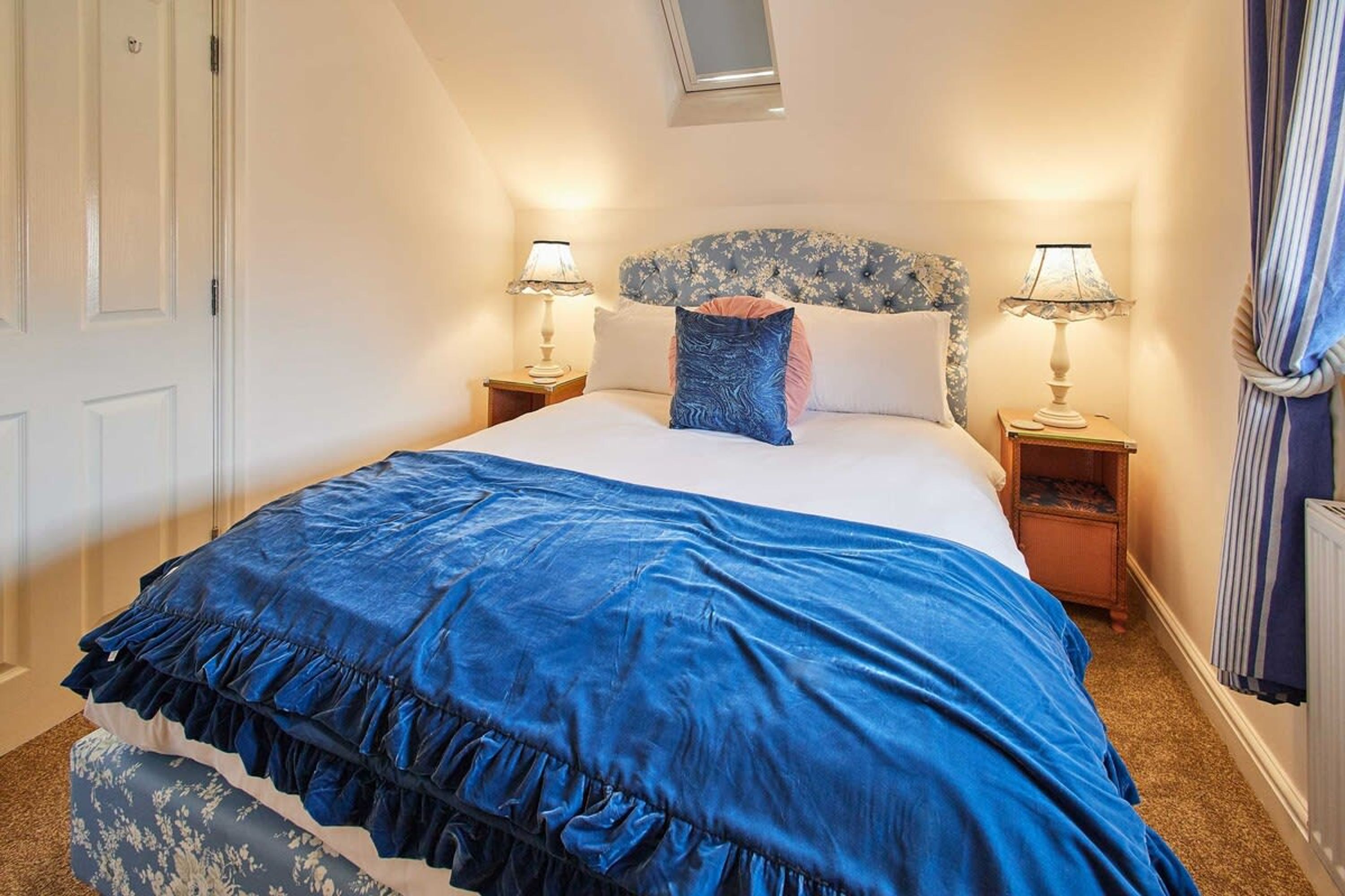 Une chambre à coucher avec un grand lit, deux tables de chevet avec des lampes, une tête de lit ornée d’un motif floral et une fenêtre avec des rideaux.