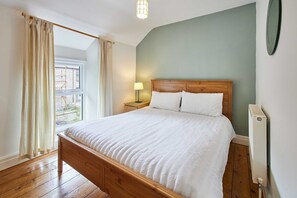 A bedroom with a wooden bed, a window with curtains, a bedside lamp, and a radiator. - Host Stay Trefor Cottage (Caernarfon)