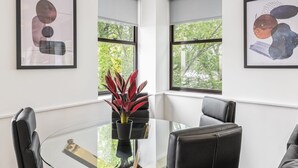 Ein moderner Essbereich mit einem Glastisch, umgeben von schwarzen Ledersesseln, abstrakte Kunstwerke an den Wänden und Blick auf Grünfläche vor dem Fenster.