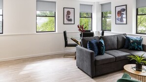 Ein modernes Wohnzimmer mit einem grauen Sofa, einem grünen Sessel und einem runden Couchtisch. Zwei Pendelleuchten, ein schwarzer Sessel und eine Pflanze auf dem Couchtisch sind ebenfalls zu sehen.
