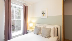 Una cama bien hecha con almohadones a rayas, una lámpara de noche y una ventana con cortinas.
