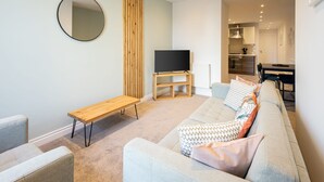 Una sala de estar moderna con un sofá, un banco de madera, un televisor sobre un mueble bajo y una cocina al fondo.