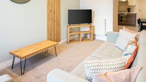 Una sala de estar moderna con un sofá, una mesa de centro, un televisor sobre un soporte de madera, una pared de acento de madera y una cocina al fondo.