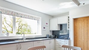 A modern kitchen with white cabinets, a wooden dining table, and a window with a view of trees. - Host Stay Manifold (Northallerton)