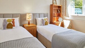 A hotel room with two beds, a wooden nightstand with a lamp, a bookshelf, and a window with a view of greenery. - Host Stay Manifold (Northallerton)