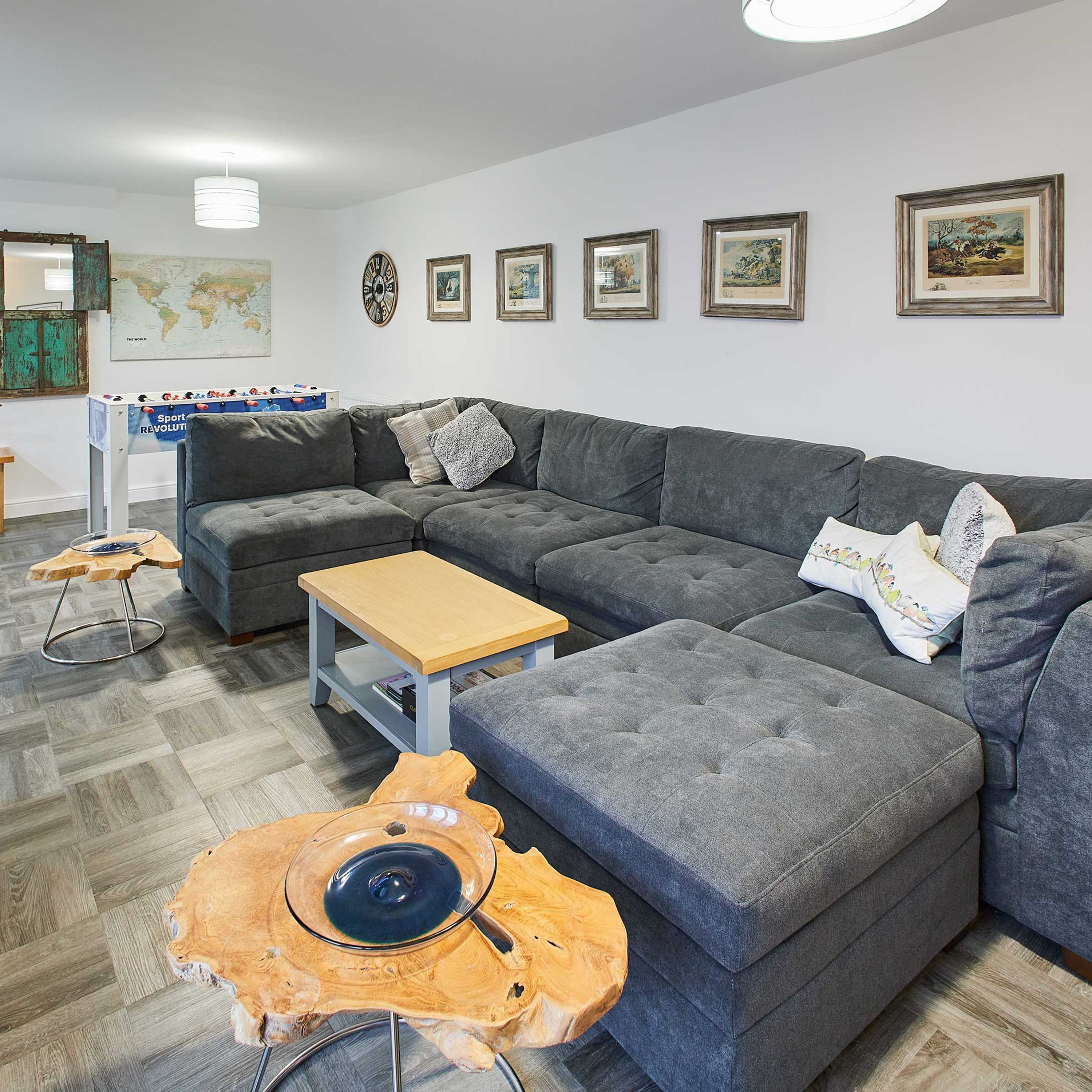 Un salon moderne avec un canapé d’angle gris, une table basse en bois et une table basse avec un plateau en verre. Des tableaux encadrés ornent le mur, ainsi qu’une carte du monde.