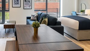 A modern, well-lit room with a large bed, a sofa, a dining table, and a small desk. There are large windows with a view of a brick building outside. - Host Stay Richmond Place 003 (Liverpool)