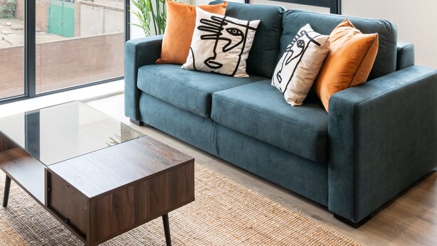 Una sala de estar moderna con un sofá azul, una mesa de centro de madera y un ventanal que ofrece vistas a un edificio y un portón verde.