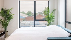 Un dormitorio moderno con una cama grande, dos sillas, un escritorio y vistas a edificios a través de un ventanal amplio.