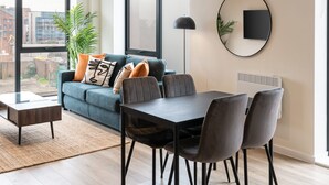 Una sala de estar moderna con sofá, una mesa de comedor con sillas, una lámpara de pie y un ventanal con vistas a edificios.