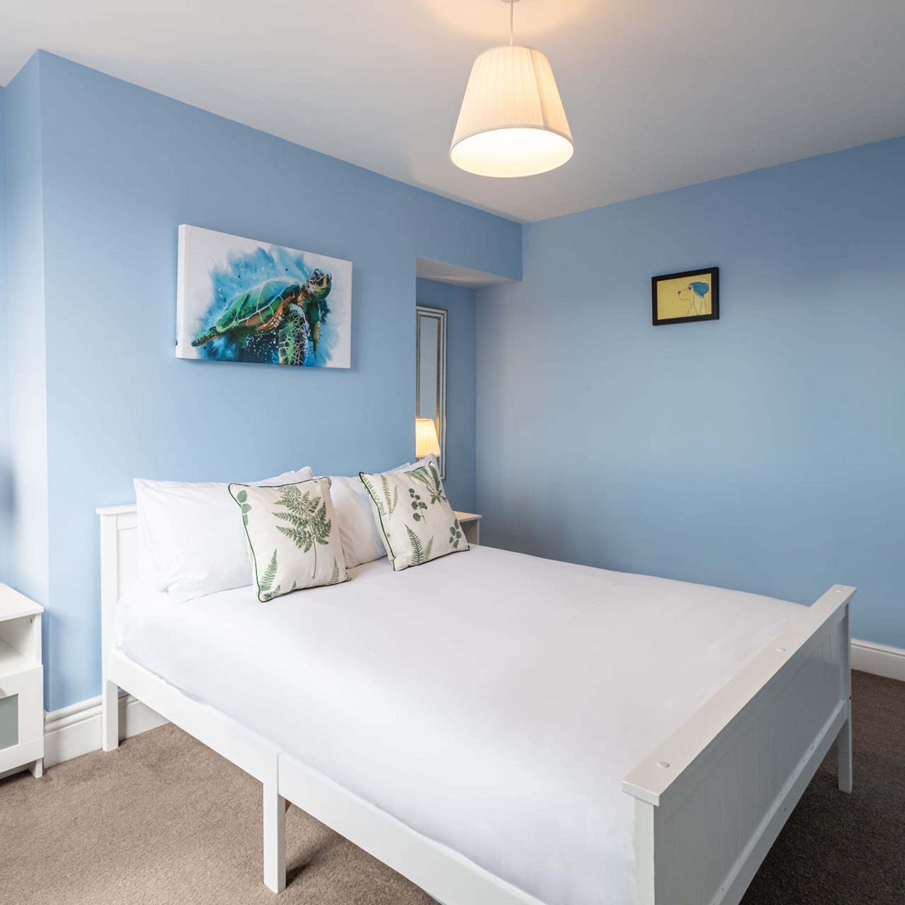 A bedroom with a white bed, blue walls, a ceiling light, a framed picture, and a bedside table.