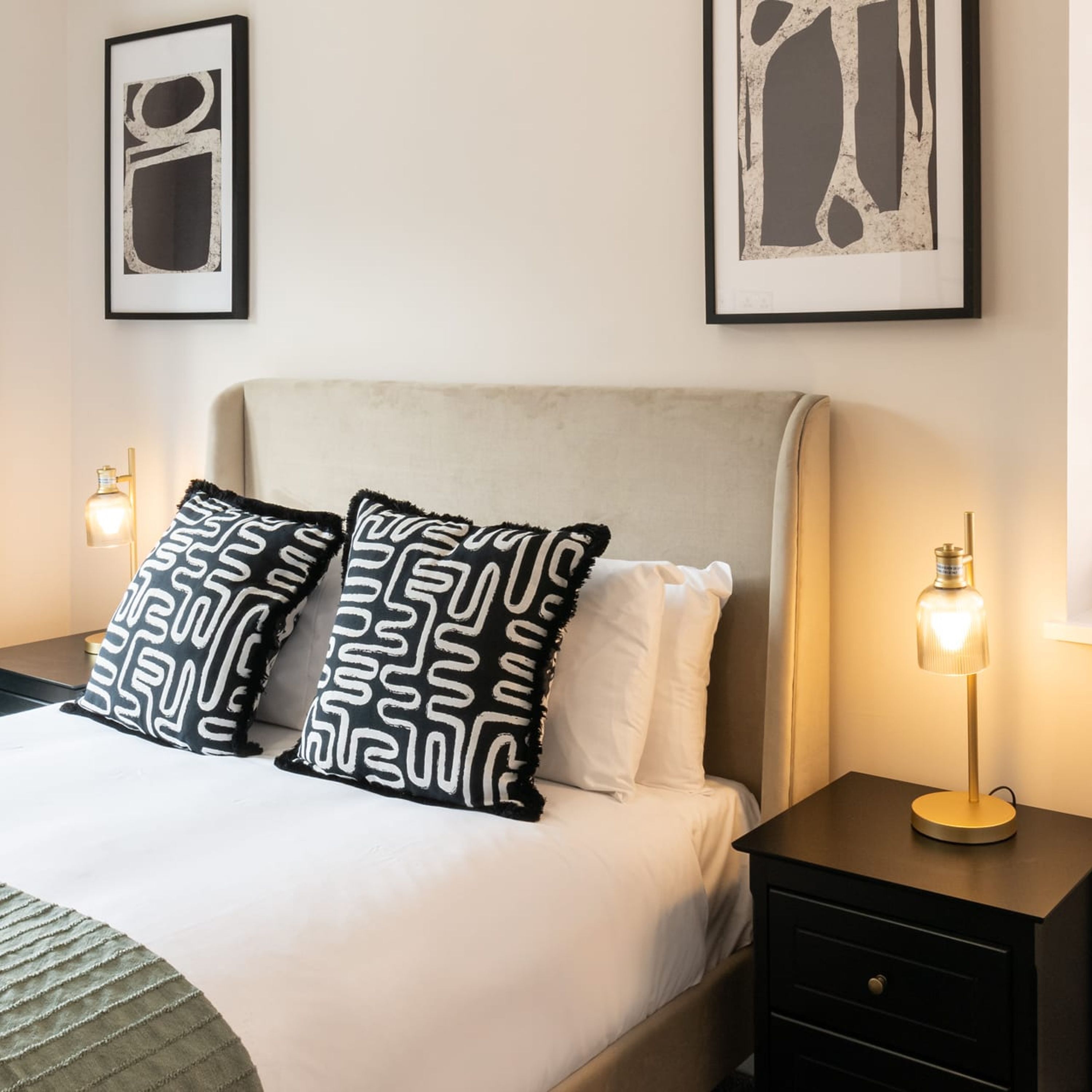A neatly made bed with patterned pillows, a bedside table with a lamp, and two framed artworks on the wall.