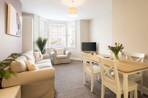 A living room with a beige sofa, a white armchair, a wooden dining table with four chairs, a flat-screen TV, and a window with blinds. - Host Stay Sandy Bay (Scarborough)