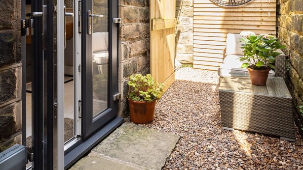 A small courtyard with a wooden fence, a stone wall, a large clock, and potted plants. - Host Stay Rock End (Matlock)
