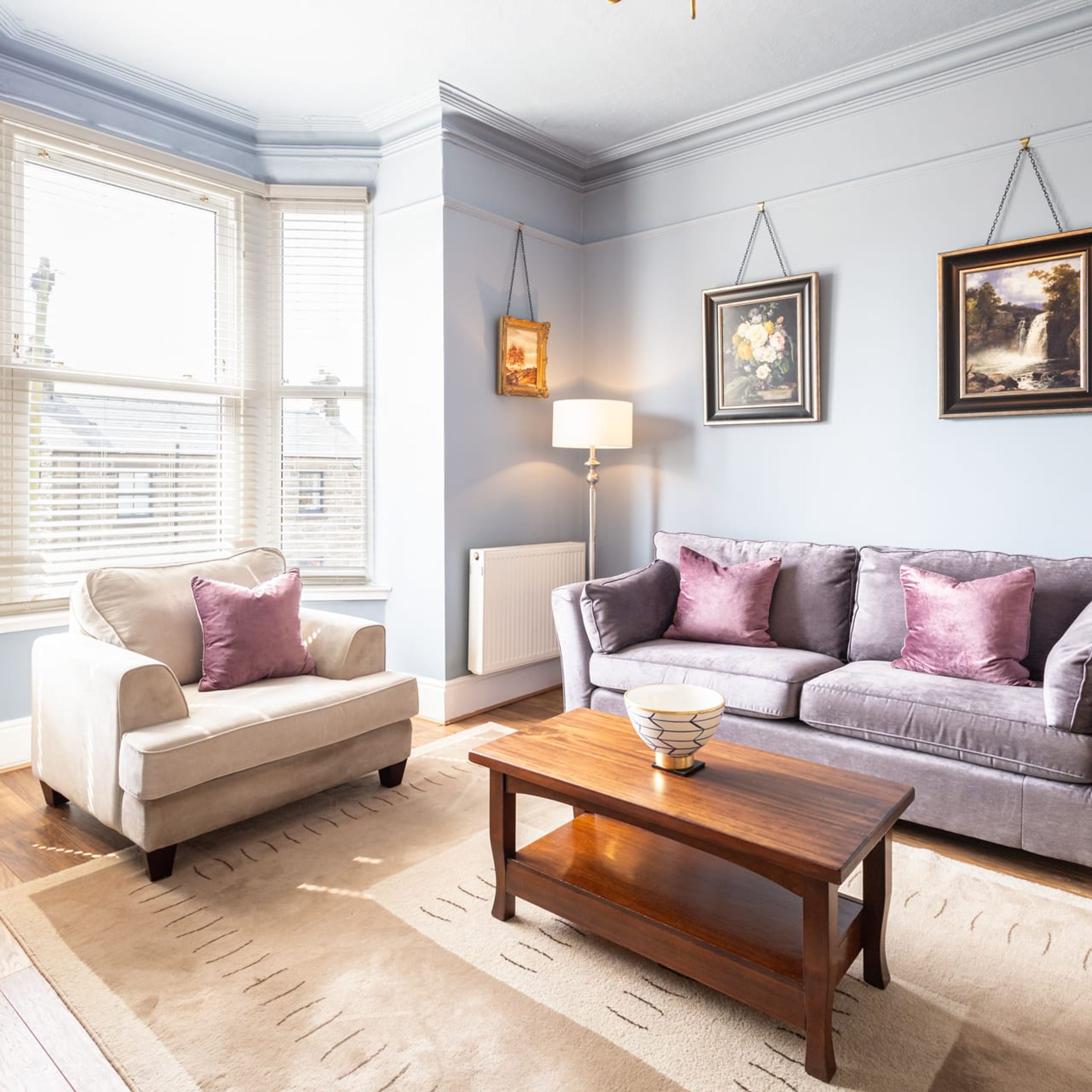 A living room with a beige sofa, a grey sofa, and a wooden coffee table. There are two framed pictures on the wall and a floor lamp.