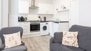 A modern kitchen with white cabinets, a stainless steel range hood, and a built-in oven. There are two grey sofas with decorative pillows, a washing machine, and a small coffee station. - Host Stay North Sands Rest (Scarborough)