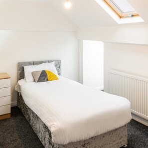 Un dormitorio con cama, mesita de noche, cómoda, radiador y tragaluz.