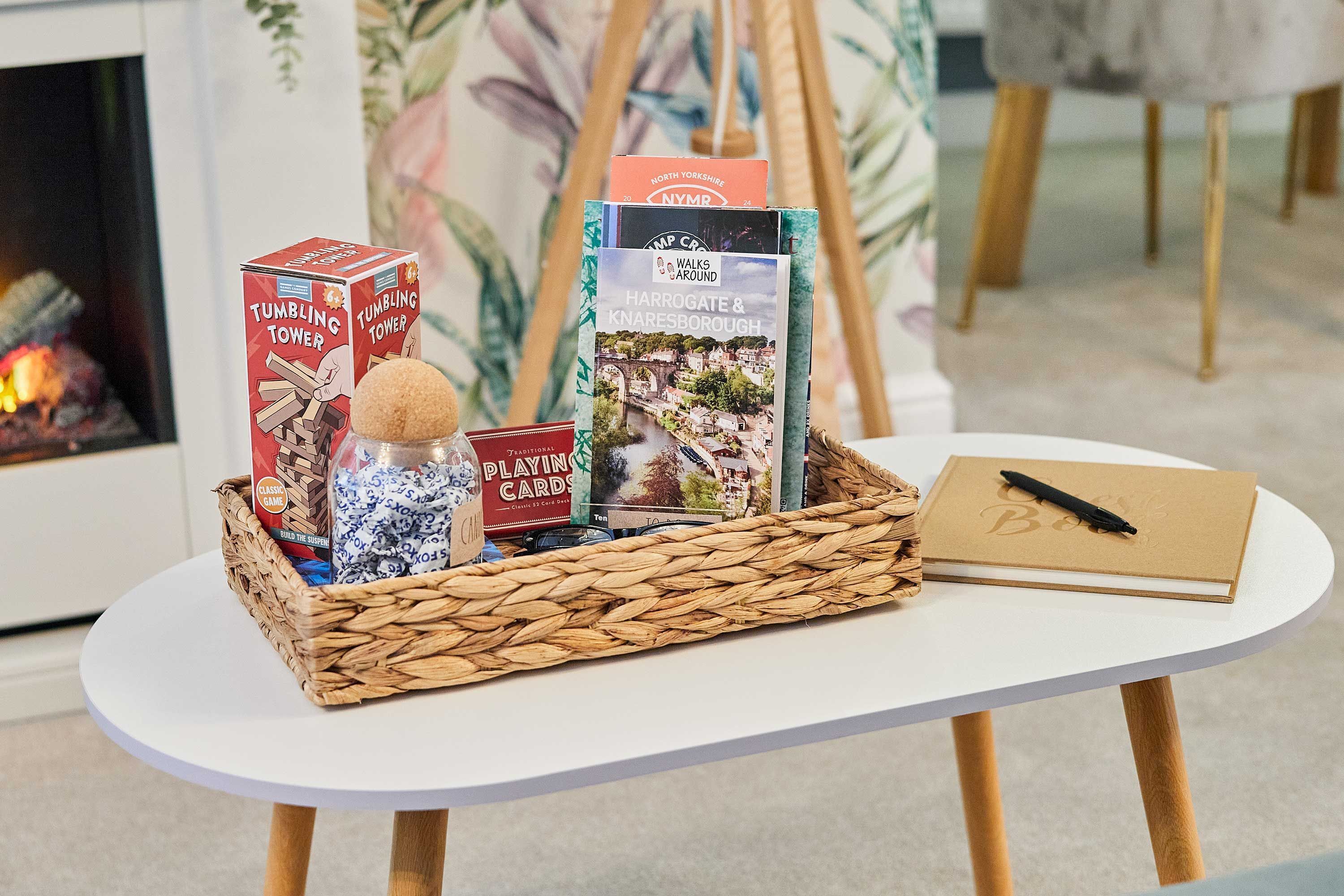A table with a woven basket containing a box labeled "Tumbling Tower," a bottle of honey, a box of playing cards, and a notebook with a pen.